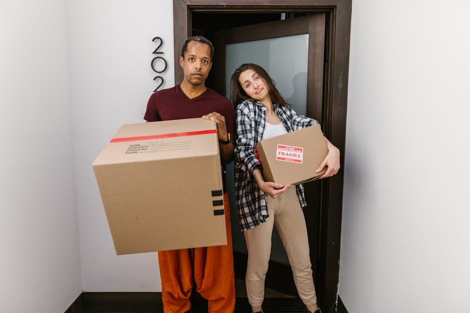 A man and a woman standing inside a doorway during a home relocation process, both holding cardboard boxes. The man, wearing an orange pair of trousers and a maroon T-shirt, holds a large box with red and black markings indicating fragile contents. The woman, dressed in beige trousers and a checked shirt, carries a smaller cardboard box with a red 'Fragile' label. They are positioned on a narrow corridor with a light grey wall on the left and a dark wooden door frame behind them, suggesting the interior of a residential property. The image captures the loading or unpacking stage of furniture transport, typical of professional removals services like those provided by Man and a Van Lambeth. The environment is well lit, emphasizing the logistical activity involved in house removals and packing. This scene illustrates the careful handling and organization during furniture transport and moving processes, highlighting the professionals involved in home relocation tasks.
