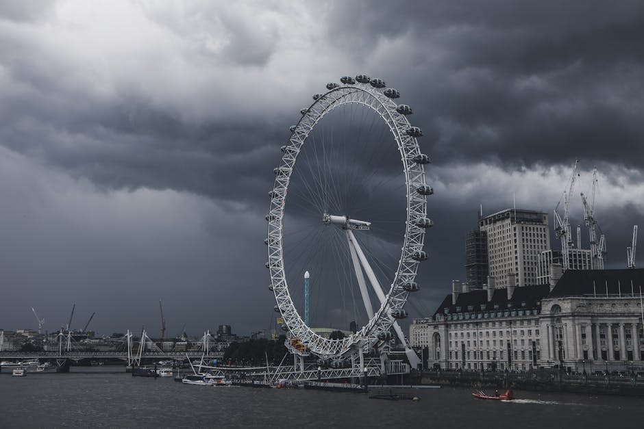 A large observation wheel, known as the London Eye, is situated along the River Thames under a dark, cloudy sky. The wheel is supported by a white A-frame structure and has multiple enclosed passenger capsules attached around its rim. Several construction cranes and tall buildings are visible in the background, indicating an urban environment. The river below features boats and piers, with some docked near the riverbank. This scene reflects a prominent London landmark, often seen in cityscapes associated with house removals and relocation services, highlighting the importance of transport logistics in home moving processes, especially in areas with iconic structures like the London Eye. The overcast weather adds a dramatic atmosphere to the scene, which is relevant for visual representations of moving and furniture transport in London.