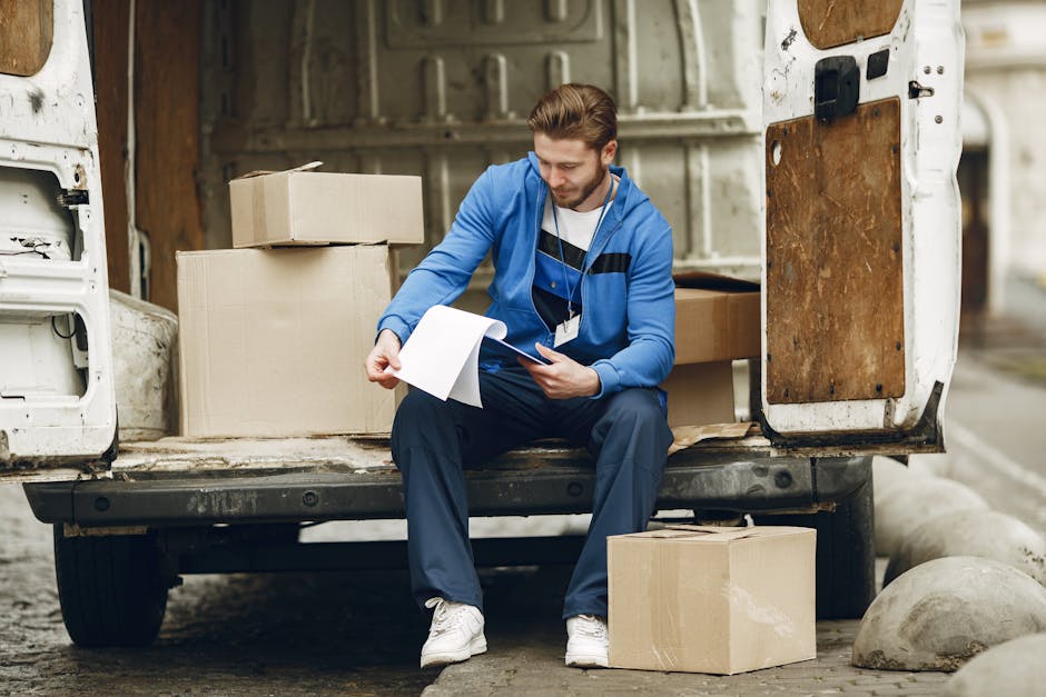 A man wearing a blue jacket, dark trousers, and white sneakers is seated on the edge of an open rear door of a white moving van, surrounded by cardboard boxes and packing materials. He is holding and reviewing a clipboard with papers, possibly checking a packing list or inventory. The van's interior shows additional cardboard boxes stacked inside, some wrapped in protective plastic or fabric, ready for home relocation. The scene takes place outdoors on a street with large concrete bollards visible on the pavement, and the van appears to be in the process of loadings or unloadings furniture and boxes. Man and a Van Lambeth is involved in furniture transport and moving logistics, with visible equipment such as trolleys and straps in the background, supporting efficient packing and furniture transport during the relocation process.
