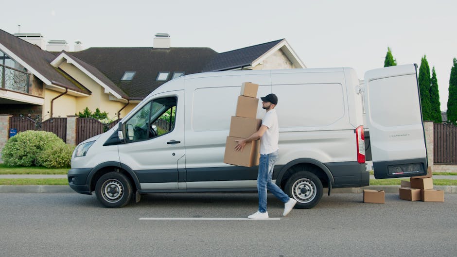 A man dressed in casual clothing, including a white t-shirt, blue jeans, and a black cap, is seen lifting a medium-sized cardboard box while standing next to an open side door of a white panel van parked on a residential street. The van's rear door is partially open, revealing additional cardboard boxes inside, some stacked on top of each other. Several other cardboard boxes are placed on the pavement nearby, indicating a home relocation or furniture transport process. Behind the man and the van, there are modern residential houses with pitched roofs, chimney stacks, and a well-maintained garden fence with greenery. The scene suggests an active loading process during a house removals service offered by Man and a Van Lambeth, focusing on packing and moving logistics. The outdoor environment is well-lit, with clear skies, emphasizing the practical aspects of moving services, including furniture transport and packing activities.