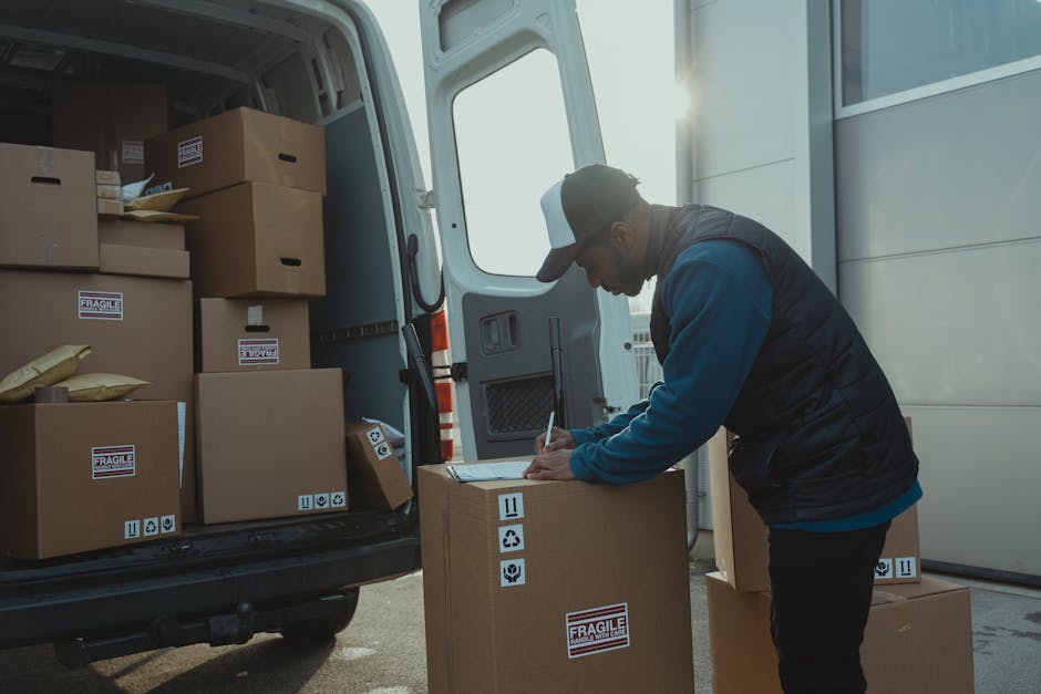 A man wearing a blue jacket, dark trousers, and white sneakers is seated on the edge of an open rear door of a white moving van, surrounded by cardboard boxes and packing materials. He is holding and reviewing a clipboard with papers, possibly checking a packing list or inventory. The van's interior shows additional cardboard boxes stacked inside, some wrapped in protective plastic or fabric, ready for home relocation. The scene takes place outdoors on a street with large concrete bollards visible on the pavement, and the van appears to be in the process of loadings or unloadings furniture and boxes. Man and a Van Lambeth is involved in furniture transport and moving logistics, with visible equipment such as trolleys and straps in the background, supporting efficient packing and furniture transport during the relocation process.