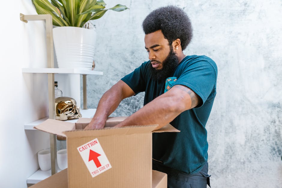 A man with an Afro hairstyle and a beard is seen packing items into a cardboard box during a home relocation process at a residential property. He is wearing a dark blue T-shirt and is standing inside a room with a light grey textured wall. Behind him, there is a white metal shelf holding a large white pot with a green leafy plant, a small decorative gold skull, and white ceramic cups. The man is focused on placing items into the box, which has a red and white caution sticker indicating the side that should face up. This staged packing activity, involving careful handling of belongings, is part of the furniture transport and packing and moving tasks managed by Man and a Van Lambeth, a professional removal service covering house removals and relocation logistics.