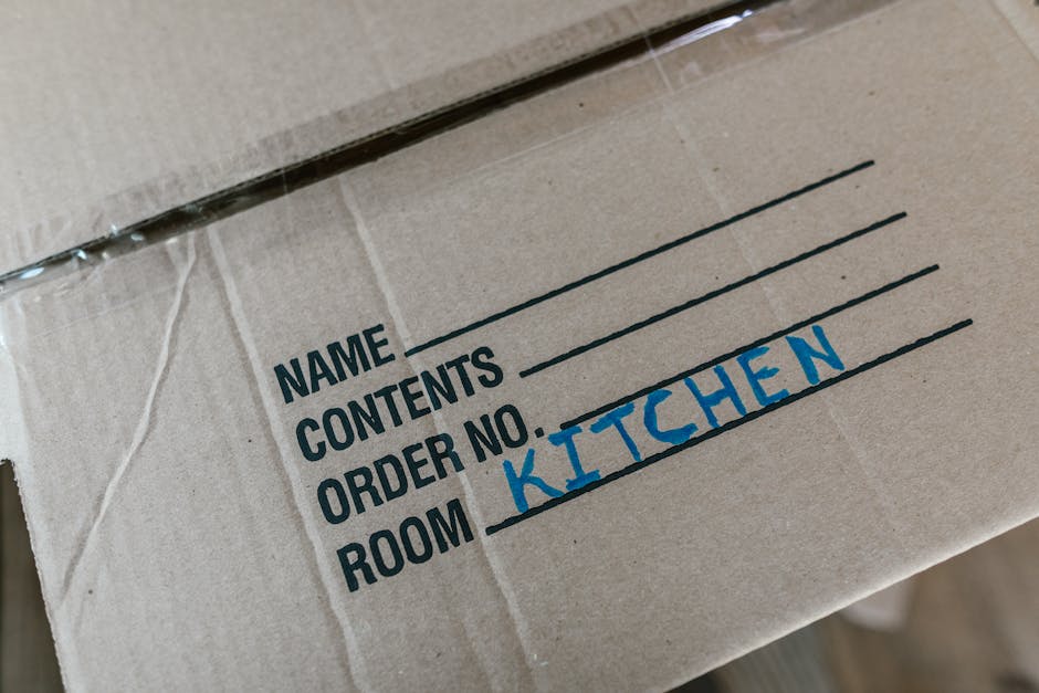 Close-up image of a brown cardboard moving box with a black printed label listing fields for name, contents, order number, and room. The 'Room' field is handwritten in blue marker, with 'KITCHEN' written inside it. The box is partially sealed with clear tape, indicating packaging for home relocation. The background suggests an indoor setting, possibly a hallway or near an entrance, where house removals and packing processes are taking place, consistent with services provided by Man and a Van Lambeth. The focus on the labelled box highlights organisation and preparation during furniture transport and packing during a house move.