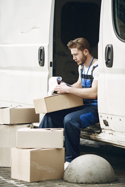 A man dressed in work overalls sitting on the edge of a white moving van's open side door, surrounded by stacked cardboard boxes of various sizes, some placed on the ground and others being held. The man is reading a document, possibly a moving checklist or inventory, while preparing for a home relocation. The van is parked on a paved surface with a concrete bollard nearby, and the interior of the van appears dark inside. The scene captures the process of packing and organizing items for furniture transport and house removals, with the focus on careful planning and logistics during the loading process. The environment is well-lit, indicating daytime, and the focus is on efficient packing and vehicle loading as part of a professional moving service, such as that provided by Man and a Van Lambeth.