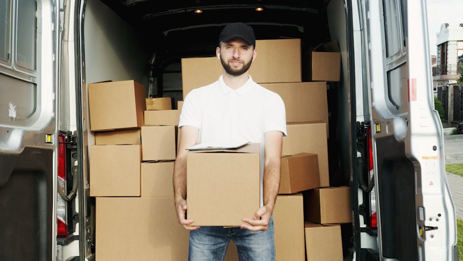 A man wearing a white shirt and black cap is standing inside the open back of a large white moving van, holding a cardboard box. The van is filled with various cardboard boxes of different sizes, some stacked on top of each other, ready for home relocation or furniture transport. The interior of the van shows packaging materials like plastic wrap and blankets used to protect furniture during transport. The environment outside appears to be a residential area, with a pathway and residential buildings visible in the background, indicating the loading process for a house move. The scene is well-lit, capturing the key elements involved in packing and moving services provided by Man and a Van Lambeth, emphasizing the logistics and handling of packing materials and furniture during the loading stage of a house removal.