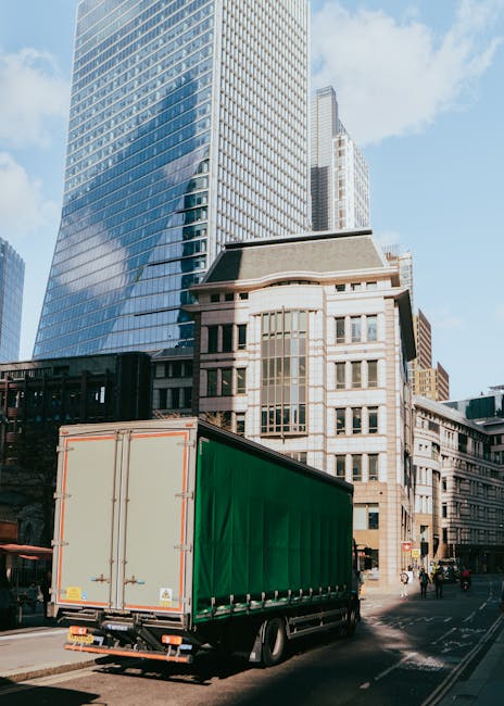 A large commercial truck with a green cargo container is parked on a city street in front of a modern glass skyscraper and older stone buildings. The truck is positioned with the rear doors facing the camera, and the vehicle is partially on the pavement and road. In the background, tall office buildings with reflective glass windows and a clear blue sky are visible. The surrounding area includes pedestrians walking along the sidewalk and street signs, indicating an urban environment. This scene relates to house removals and moving logistics, such as transportation of furniture and belongings during a home relocation, which Man and a Van Lambeth provides as part of their services in the Lambeth area.