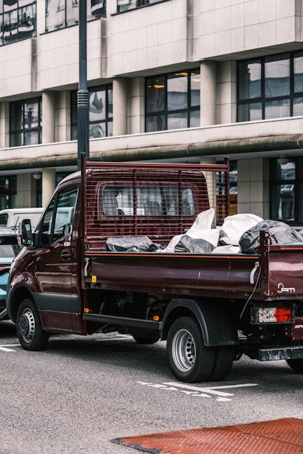 A maroon flatbed truck parked on a city street with its cargo area filled with various household items wrapped in plastic and placed within cardboard boxes, ready for a home relocation. The items include a black plastic bag and other packed belongings securely arranged on the truck bed, with protective metal rails on the sides. Behind the truck, modern office windows and a concrete building facade are visible, indicating an urban environment suitable for house removals or furniture transport services. The scene captures part of the loading process or transportation phase, with the vehicle positioned adjacent to a sidewalk on a cloudy day, and a streetlamp visible in the background. Man and a Van Lambeth occasionally support such logistics operations as part of their removals services, facilitating efficient packing and moving within the Lambeth area, aligned with local parking permits and regulations.