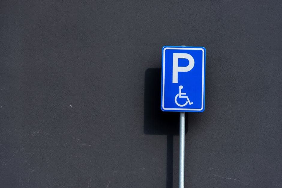 A close-up image of a blue rectangular parking sign mounted on a metal pole, positioned against a dark grey wall. The sign features a white letter 'P' and a wheelchair symbol underneath, indicating accessible parking for disabled individuals. The lighting creates a shadow of the sign on the wall behind it. The sign is located outdoors, likely near a property entrance or parking area, relevant to home relocation and furniture transport services. This visual detail supports the understanding of parking arrangements and permits required during house removals, as provided by Man and a Van Lambeth, which specializes in removals and relocation services in the Lambeth area.