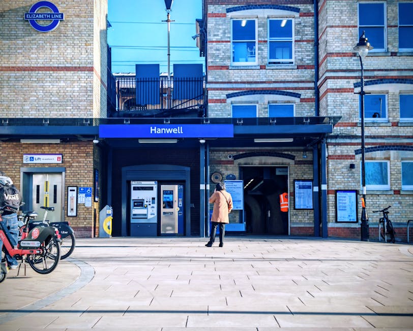 A person dressed in a beige coat and dark trousers is standing outside Hanwell Underground station, part of the Elizabeth Line, on a paved plaza. The station entrance features a black awning with a bl
