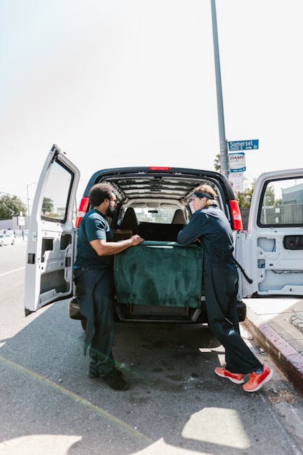 Two movers from Man and a Van Lambeth are engaged in a home relocation process, carefully loading a large, dark green piece of furniture wrapped in fabric or moving blankets into the back of a black cargo van parked on the side of a street. The interior of the van is visible with its open doors, revealing a spacious cargo area designed for furniture transport. One mover, wearing a blue shirt, is holding the furniture steady, while the other, dressed in a dark jacket and sneakers, assists with the lifting and positioning. The scene is set outdoors during daytime under bright, natural light, with a clear sky overhead and a street sign visible in the background, indicating a residential or urban area. Surrounding the vehicle are sections of pavement, with a curb and street markings nearby. This image exemplifies the careful handling and logistics involved in packing and loading household items during a professional removals service in Lambeth.