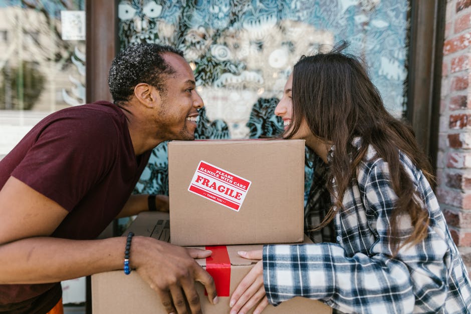Two movers from Man and a Van Lambeth are engaged in a home relocation process, carefully loading a large, dark green piece of furniture wrapped in fabric or moving blankets into the back of a black cargo van parked on the side of a street. The interior of the van is visible with its open doors, revealing a spacious cargo area designed for furniture transport. One mover, wearing a blue shirt, is holding the furniture steady, while the other, dressed in a dark jacket and sneakers, assists with the lifting and positioning. The scene is set outdoors during daytime under bright, natural light, with a clear sky overhead and a street sign visible in the background, indicating a residential or urban area. Surrounding the vehicle are sections of pavement, with a curb and street markings nearby. This image exemplifies the careful handling and logistics involved in packing and loading household items during a professional removals service in Lambeth.