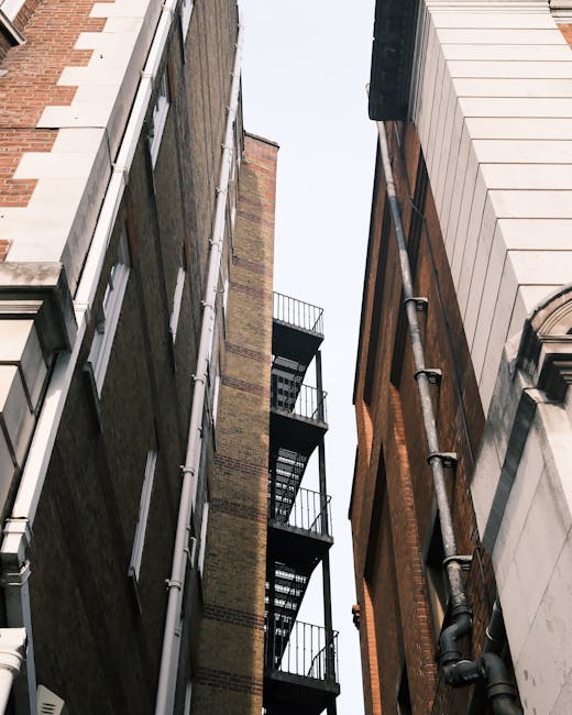 Photograph taken from ground level showing a narrow gap between two tall buildings, with fire escape staircases attached to each structure. The building on the left features a brick facade with vertical white pipes running down its side, while the building on the right has a white stone exterior with black pipes also visible. The sky above is overcast, and no vehicles or furniture are visible in the image. This setting illustrates the confined space typical of urban house relocations involving narrow access solutions, where careful planning is required for the transportation of furniture and boxes during a home removal. Man and a Van Lambeth's professional team would utilize such narrow passages to load moving containers or vans efficiently, often employing specialized equipment like trolleys and straps to navigate tight areas, ensuring safe and efficient furniture transport within complex city environments.