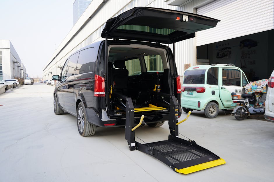 A black wheelchair-accessible van parked outside a warehouse-like building with an open rear hatch, revealing a hydraulic tail lift lowered onto the pavement for home relocation or furniture transport. Inside the vehicle, there are visible black seats, and the tail lift platform has a yellow safety edge. Surrounding the van, several smaller vehicles including a light green compact van and a scooter are parked nearby, with some wrapped packages or boxes on the scooter. The scene indicates an active loading process typical of removals services with packaging materials like cardboard and protective blankets possibly in use, supported by the professional removals company Man and a Van Lambeth. Overcast daylight provides neutral lighting for the area, conveying a scene of logistical coordination in the context of packing, moving, and transport of household belongings.